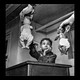 Black and white photo of two hands offering dolls to child seated at desk, child reaching for the white doll while looking at the Black doll