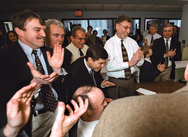 photo of reporters in crowded newsroom looking at winning announcement while smiling and applauding