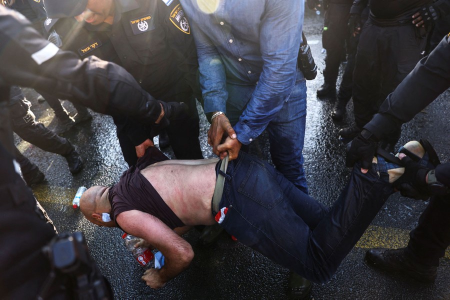 Police and security forces carry a person from a road, lifting them by their shirt, belt, and pants.