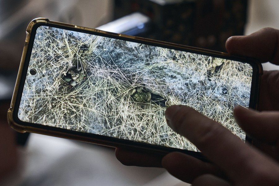 A person holds a smartphone, pointing to the screen, which shows an aerial image of soldiers in a field.