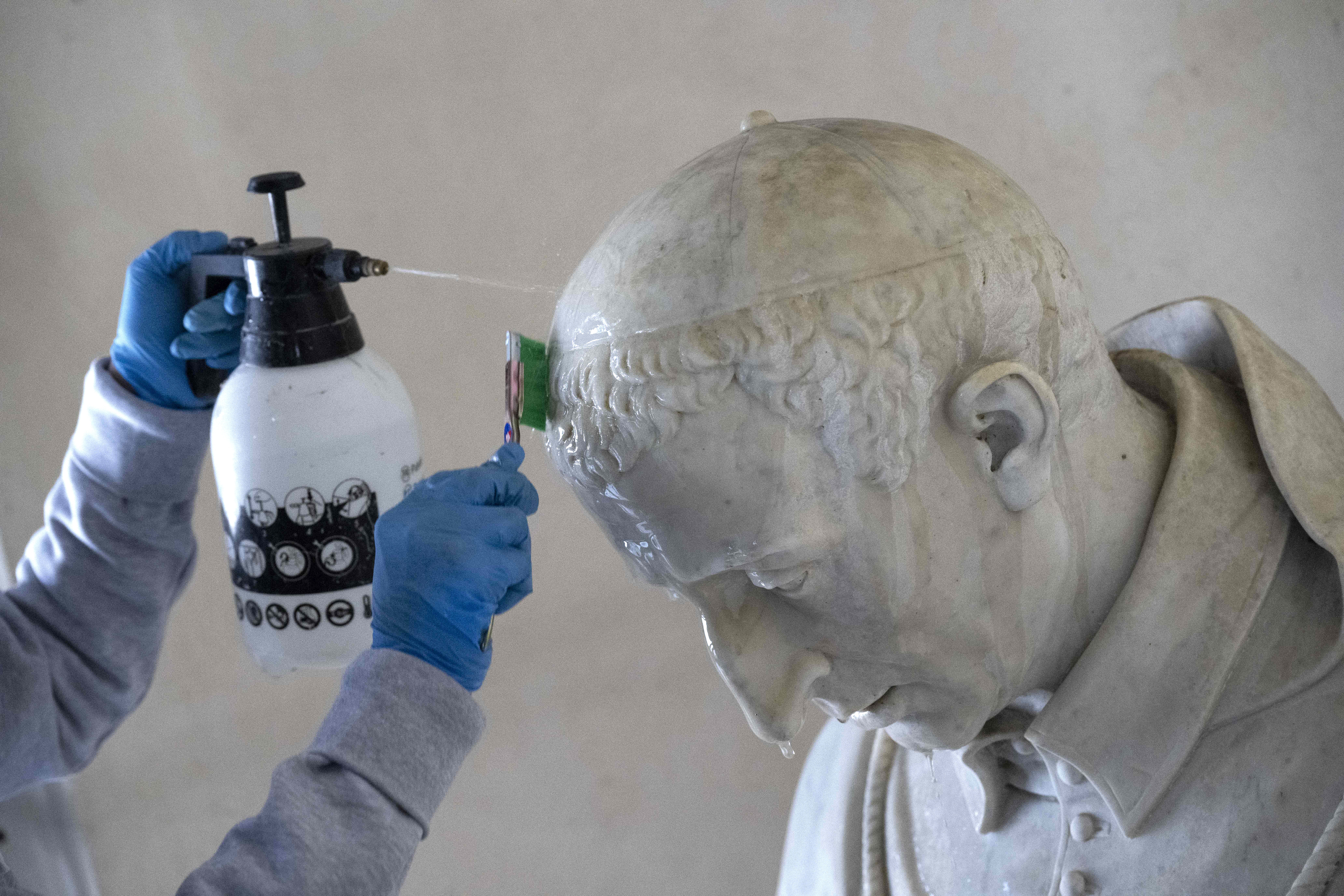 A worker sprays cleaner on the head of a statue, scrubbing with a brush.
