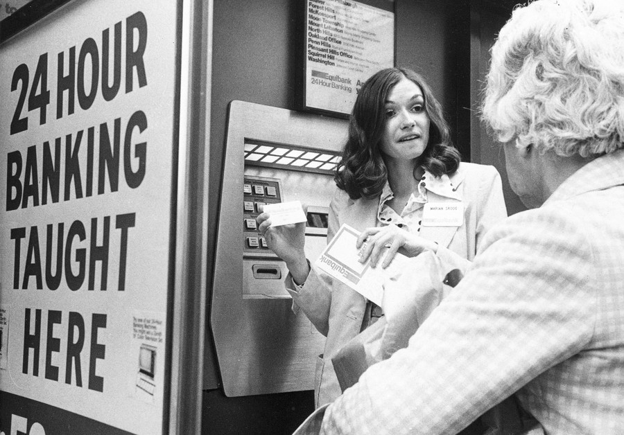 Two people stand beside an automated teller machine—one is demonstrating the machine to the other.