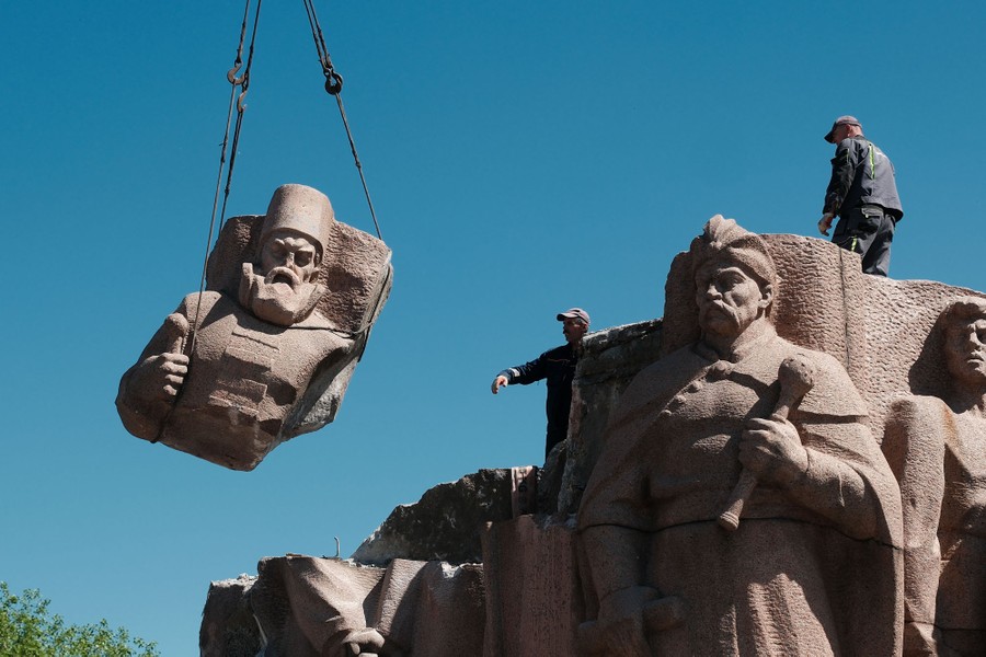 Workers dismantle a large granite group sculpture, lifting away the upper section of one figure.