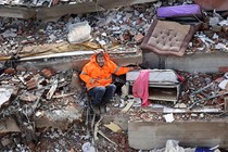 A man sits in the rubble of a collapsed building, his left arm reaching out and holding the hand of his daughter, who lies dead, trapped beneath the debris.