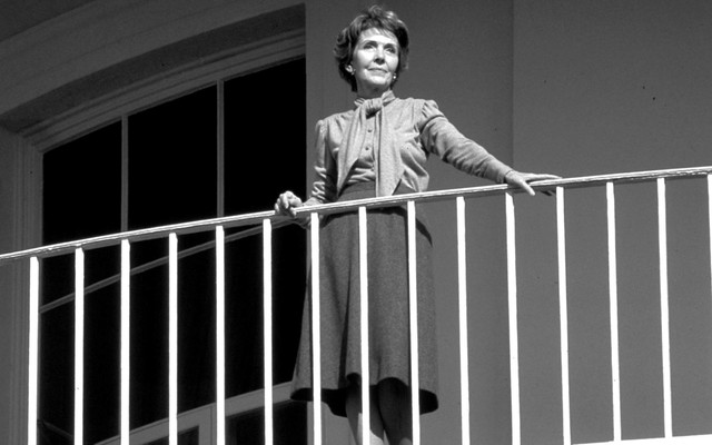Nancy Reagan stands on a balcony, watching an unseen President Ronald Reagan depart the White House.