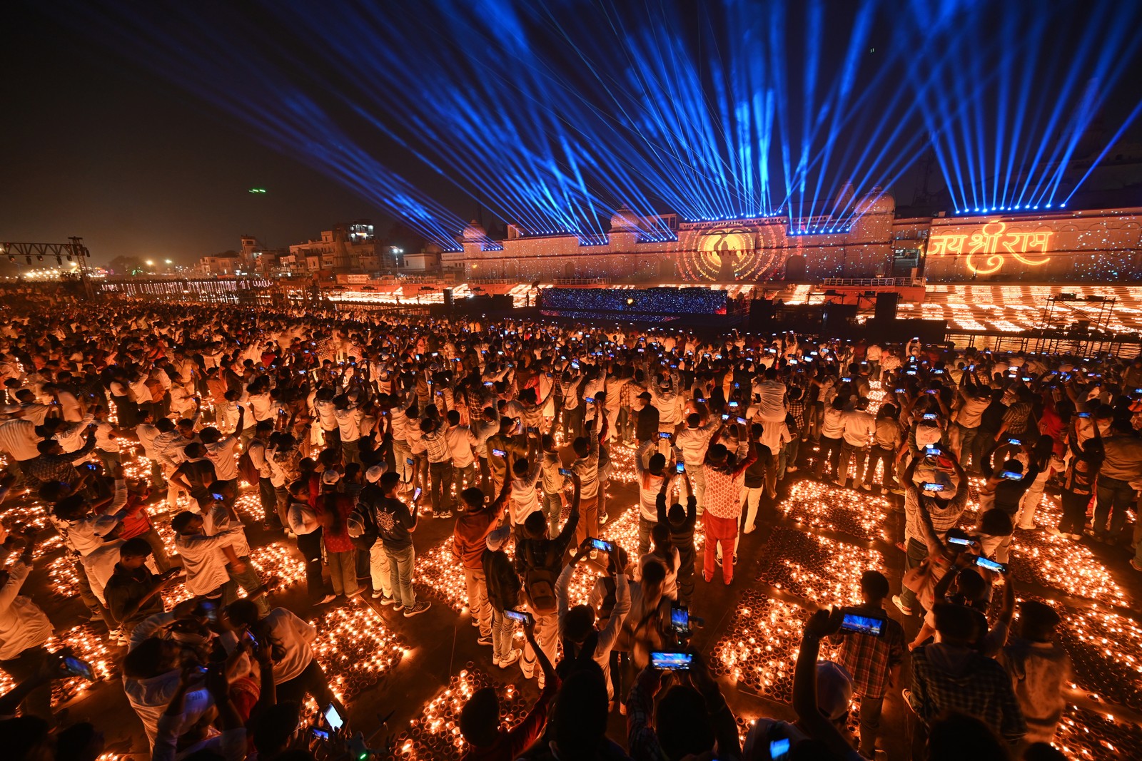 A large group of people celebrate and take photographs while standing among many lit oil lamps, and watching a light show.