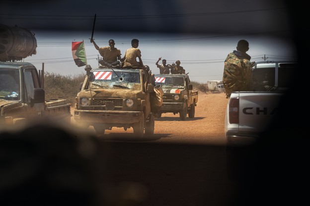 photo through car window of military vehicles and trucks driving on dirt road, one flying flag, with fighters, arms raised, sitting on tops and sides
