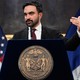 A photo of Mayor Zohran Mamdani speaking at a podium in front of an American flag and a New York City flag.