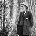 A black-and-white photo of John Muir wearing a suit and hat, standing by a large tree in a forest