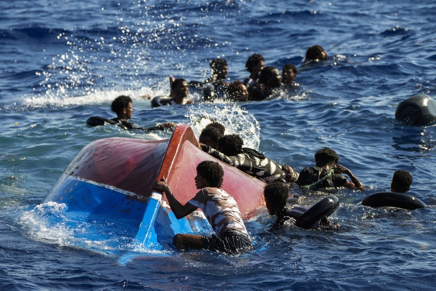 More than a dozen people swim, splash, and climb onto an overturned boat in the ocean.