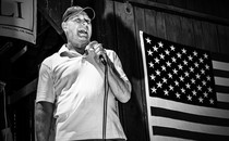 a black-and-white photo of a man speaking into a microphone next to an American flag