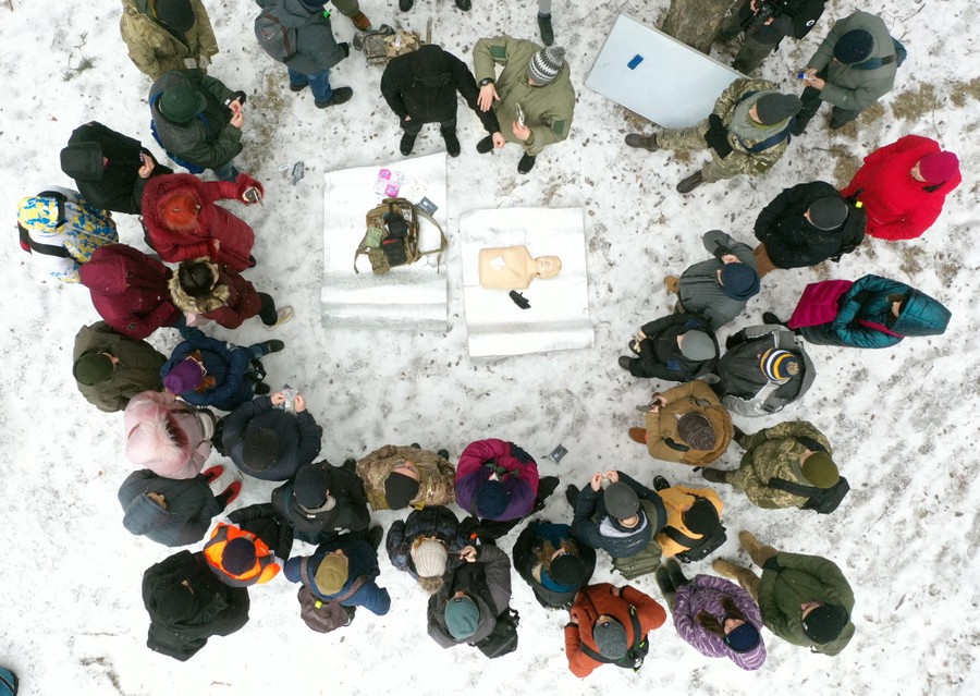 An aerial view of a couple dozen civilians standing in a circle around instructors and a CPR Doll.