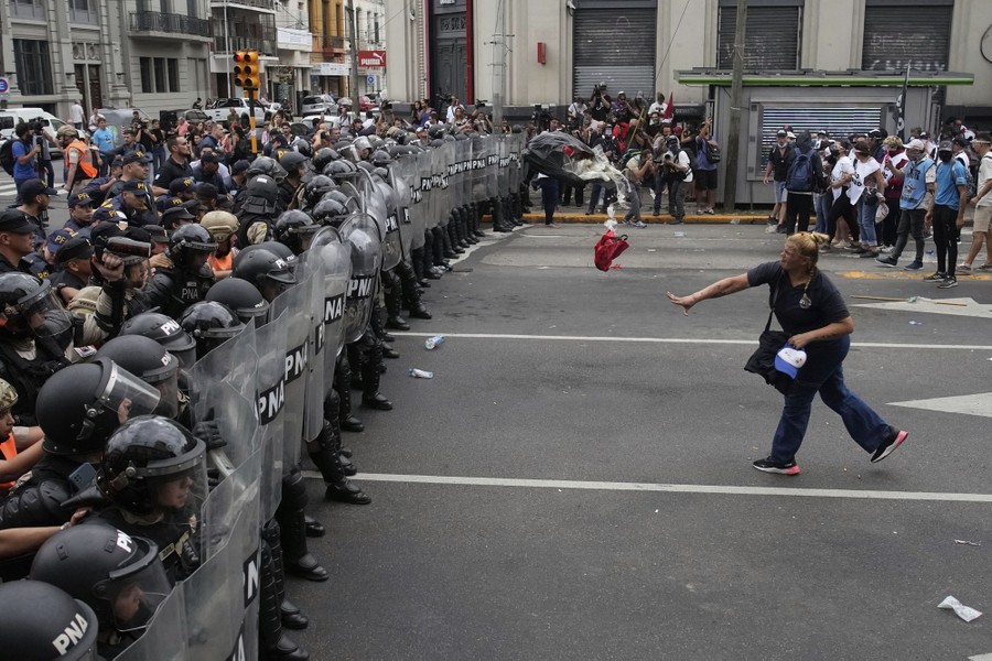 A woman throws a bag of trash at a line of riot police officers holding shields.