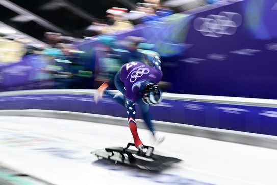 An Olympic athlete sprints on an ice track, bent over, pushing a small sled at the start of a run, legs and arms blurred by motion.