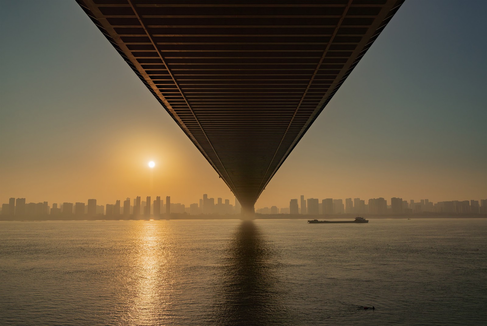 A city skyline in the distance, seen across a body of water, from beneath the deck of a huge road brisge.