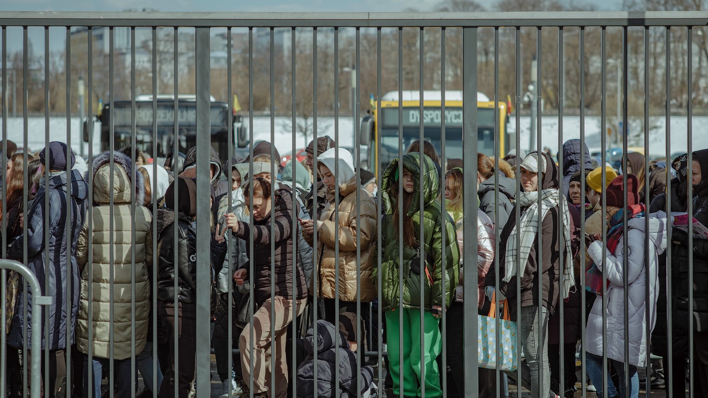 A line of people bundled up in parkas and scarves wait in a crowded line behind a tall barred metal fence with buses and snowy city in background