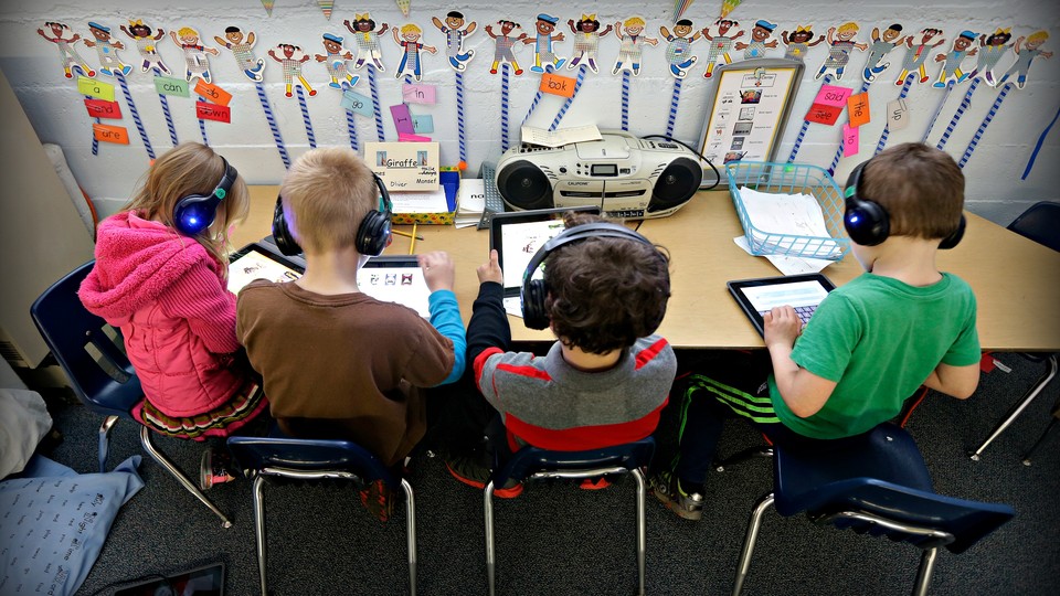 Children in a classroom use tablets while wearing headphones.