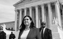 Jena Griswold, wearing a suit jacket and pearls and surrounded by men in dark sunglasses, stands in front of the Supreme Court building.
