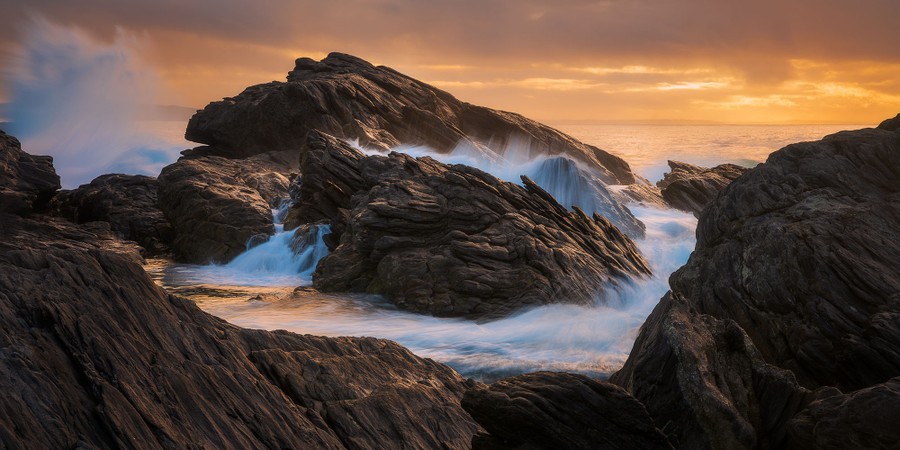 Waves crash over a rocky shoreline with the sun low in the sky.