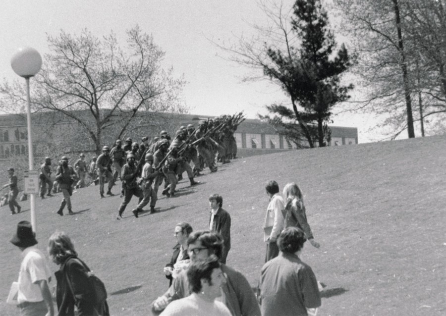 A line of armed National Guardsmen wearing gas masks marches up a hill past civilians.