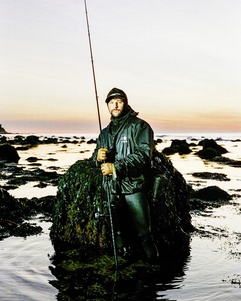 photo of man wearing wetsuit, jacket, and hat, standing in knee-deep ocean in front of waist-high rock by shore, holding long fishing pole 