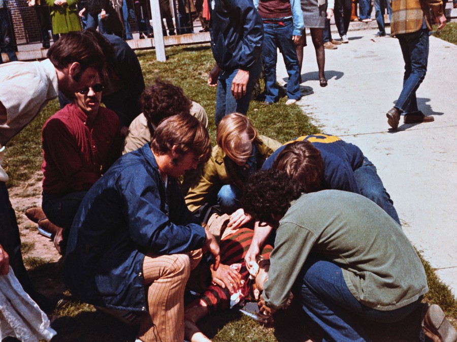 A half-dozen people kneel down to help a bleeding person lying on a patch of grass.