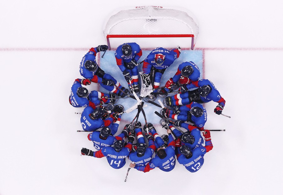 An aerial view of 14 members of a sled hockey team huddling in a circle in front of a goal.