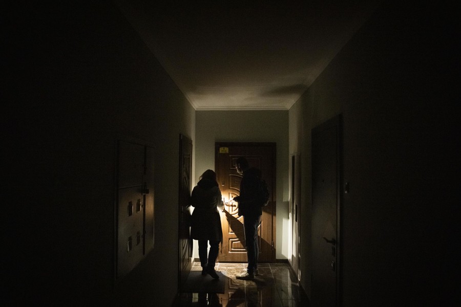 Two people stand in an apartment block hallway, at a door, lit only by a flashlight.