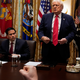 Donald Trump standing up behind a wooden table with Marco Rubio and Pete Hegseth seated beside him