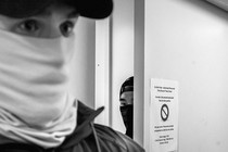 A black and white photographs of two ICE Officers, one in the foreground, and one peeking out from behind a door.