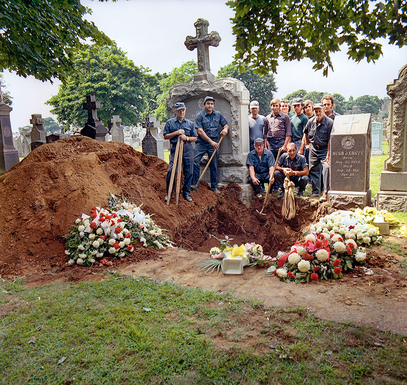 photo of group of men with shovels by hole in front of stone memorial next to large pile of dirt and funeral flowers 