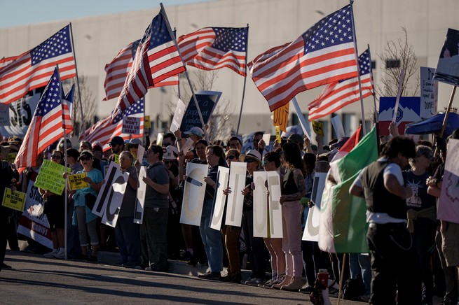 People protesting the possible new ICE detention center at the warehouse in Utah; crowd of dozens holds American flags and signs that read "ICE OUT OF UTAH"