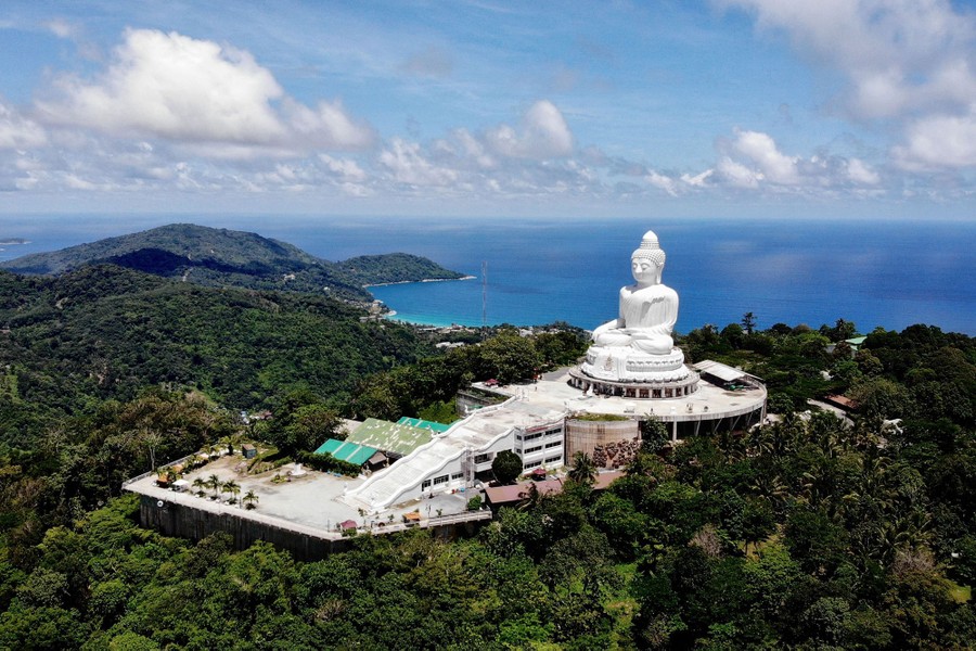 An aerial view of a giant white statue of Buddha set on a hilltop above a broad beach.