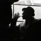 A man wearing a hat and a wool coat drinks from a glass while he sits next to a window