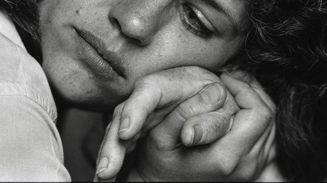 A woman with a contemplative face holds someone else's hand in hers.  His cheek rests on their hands.