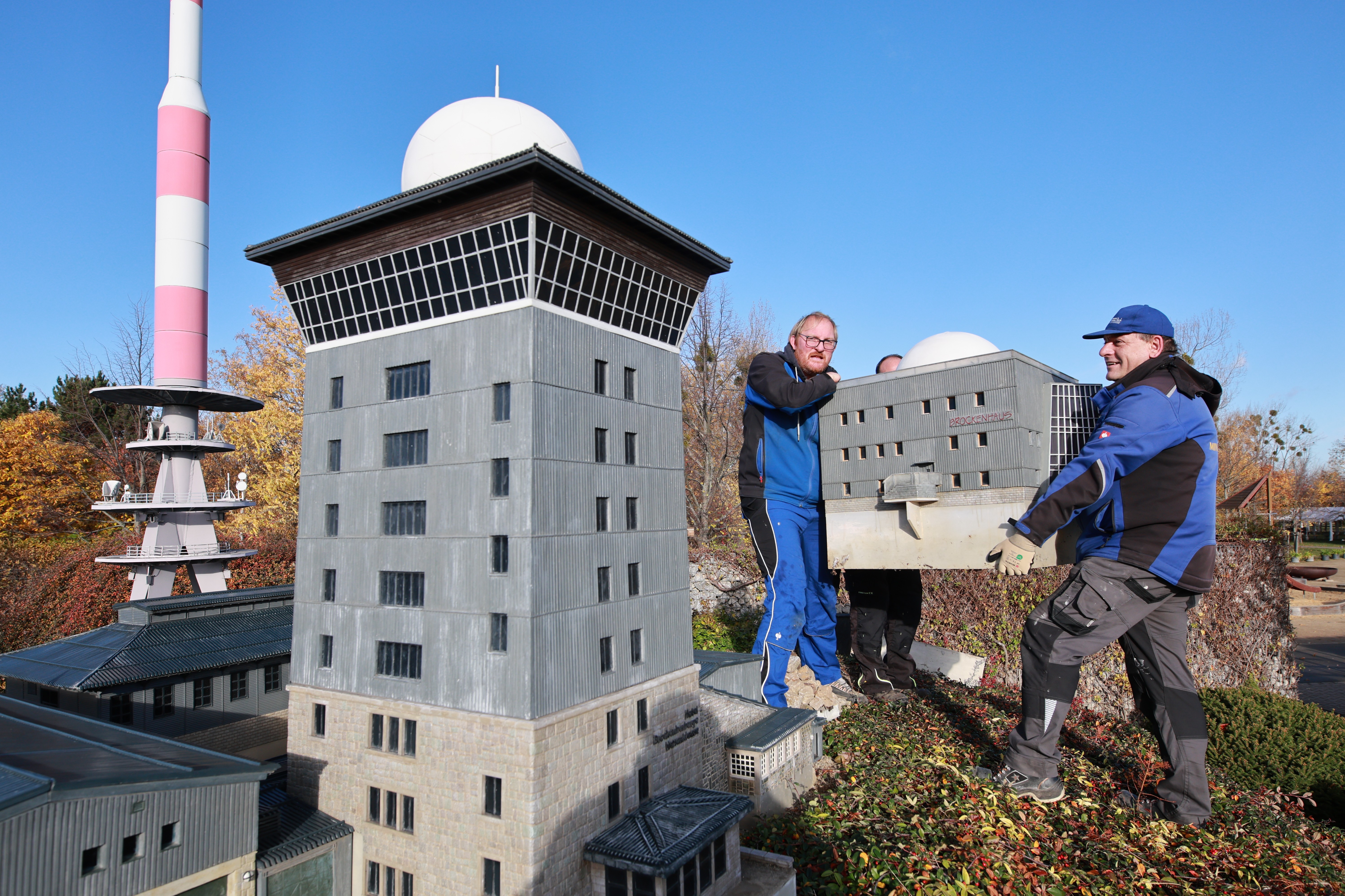 Two workers carry a scale model of a city building, beside other models.