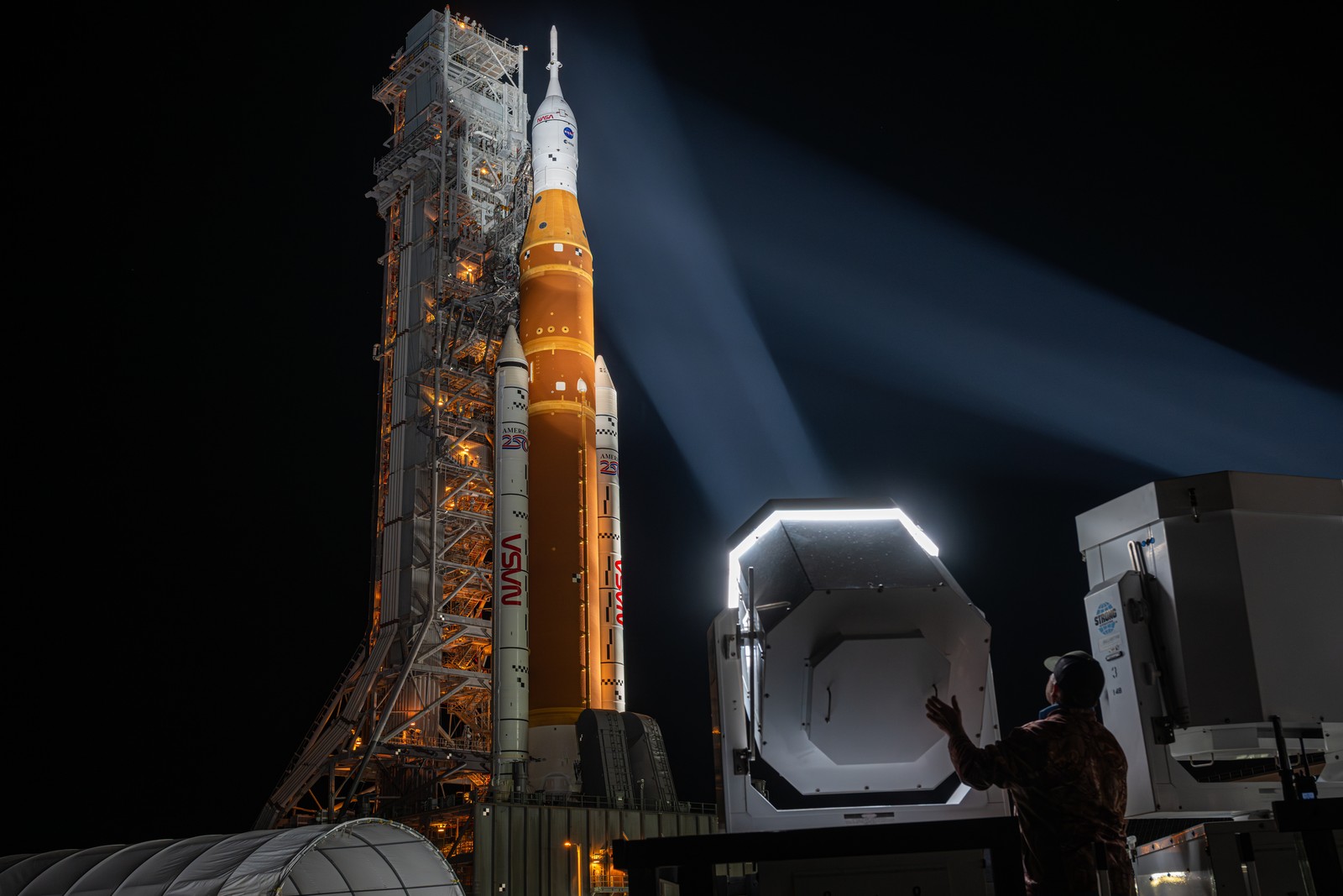 A person stands behind a spotlight that is aimed up at a tall rocket that is being rolled out to a launch pad.