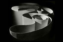 A black-and-white photo of a spotlighted spool of corrugated paper unwinding in artful shapes, surrounded by darkness.