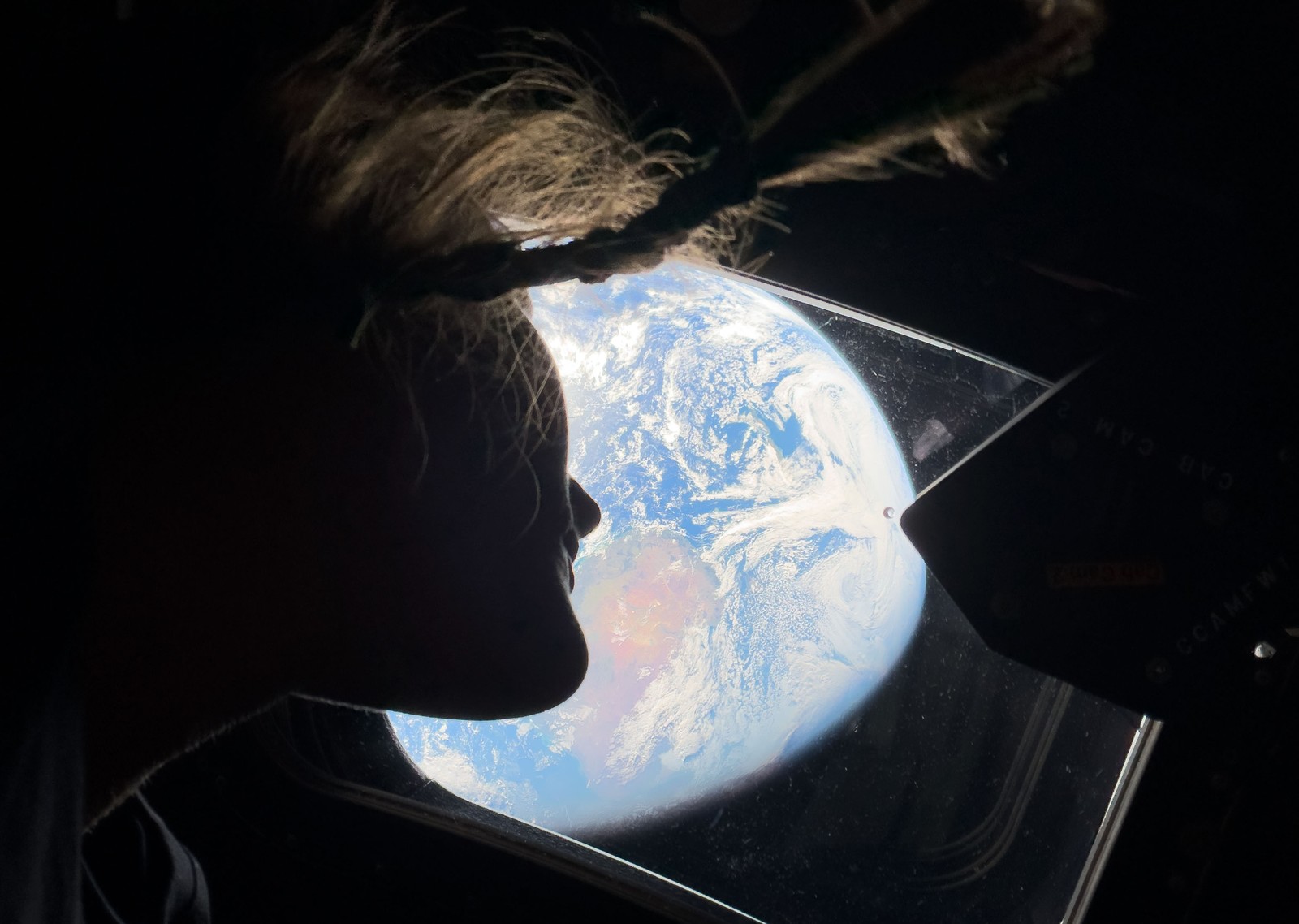 An astronaut, seen in profile, with the brightly-lit Earth in the background, seen through a window