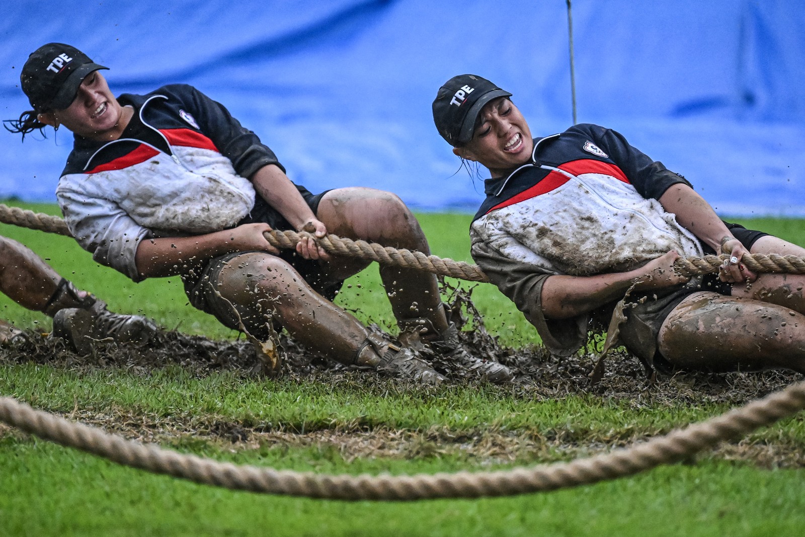 Several people strain, leaning back and pulling a rope during a tug-of-war in a muddy and grassy field.