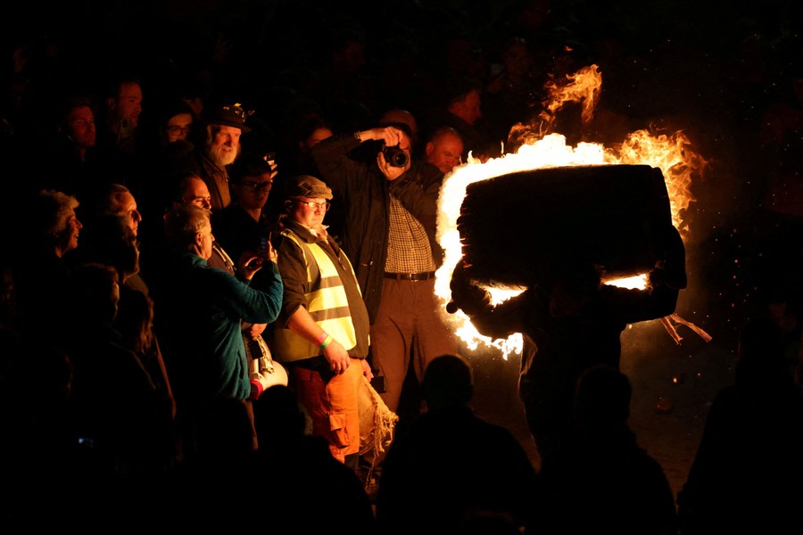 A crowd watches as a person runs past carrying a flaming barrel on their shoulders.