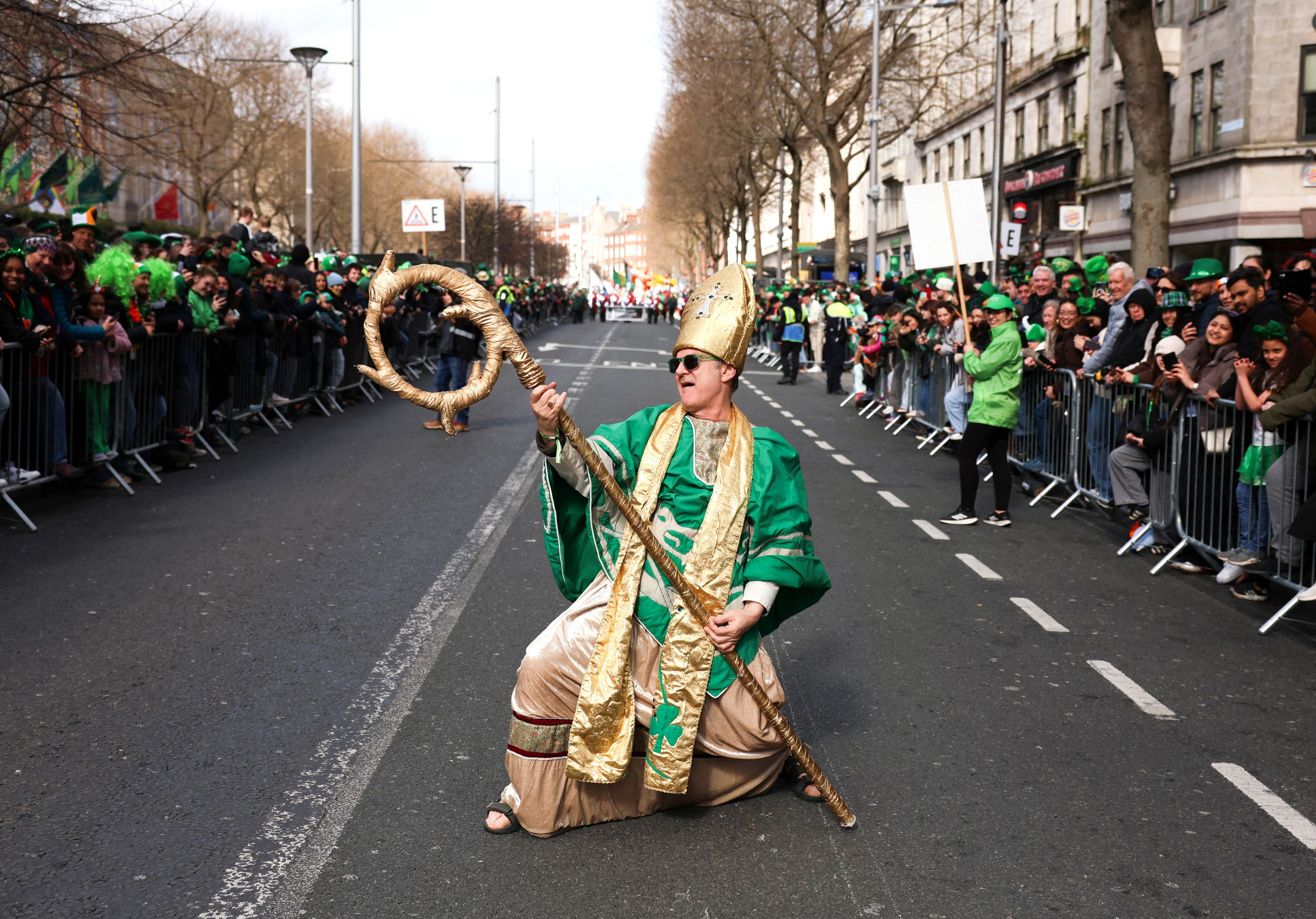A performer dressed as as Saint Patrick poses with his staff as if it were a rock guitar, during a St. Patrick's Day parade.