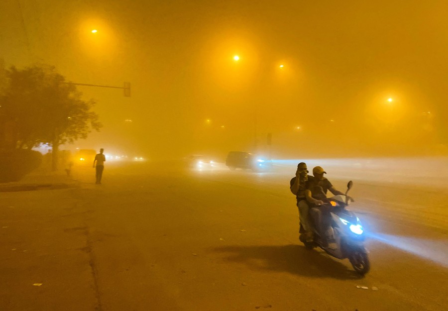 Two people ride a scooter on a city street under an orange-colored sky, thick with dust.