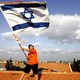 A person waving a large Israeli flag while standing on a roof with a big sky in the background