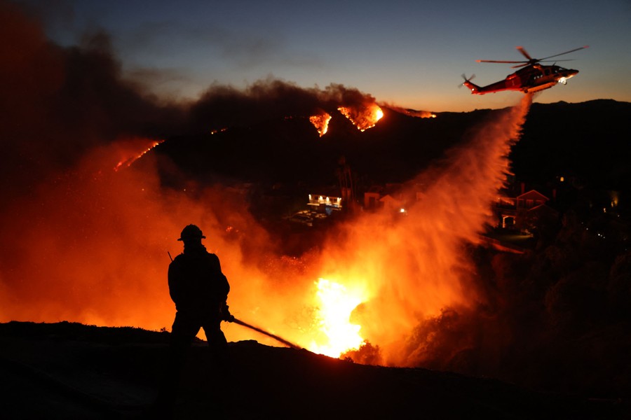 A firefighter silhouetted against a wildfire as a helicopter drops water nearby.