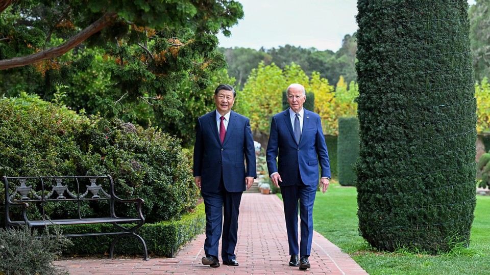 Xi Jinping and Joe Biden, both wearing blue suits, walking side by side on a brick path through a garden