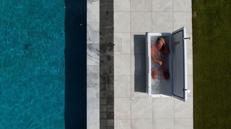 A person relaxes in a tub of ice water set on pavement beside a swimming pool.