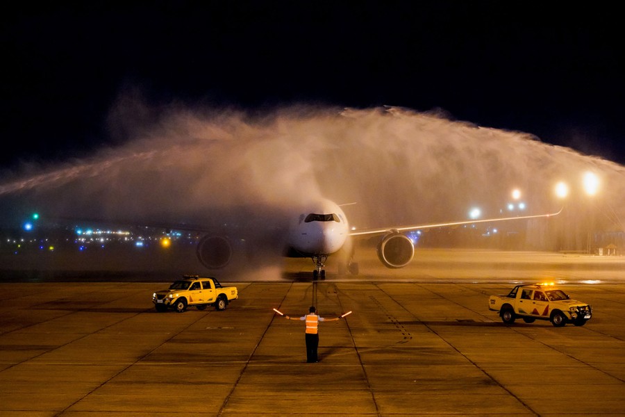 A large aircraft rolls on tarmac, showered with water from two firehoses.