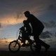 A man guides a child riding a two-wheel bike. It's sunset, and the figures appear as silhouettes.