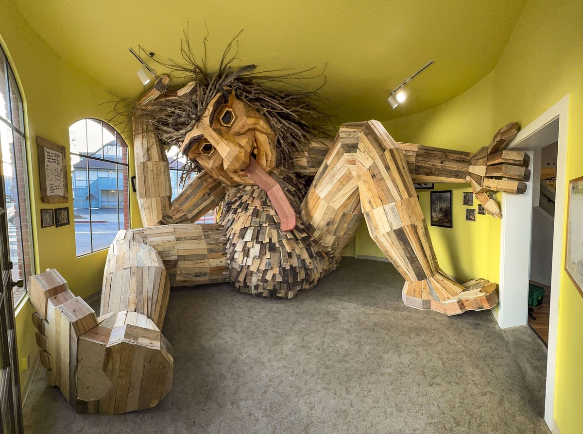 A large wooden troll sculpture sits inside a room, looking cramped, its head against the ceiling.